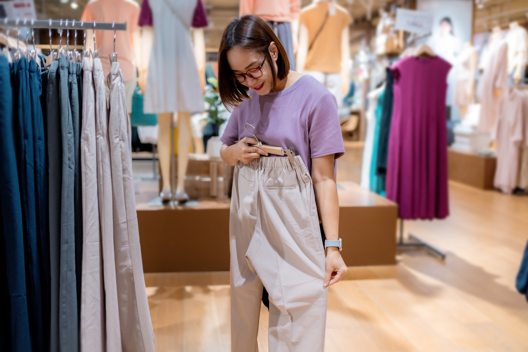 Young woman choosing pants in a fashion store, holding beige trousers near waist, comparing size